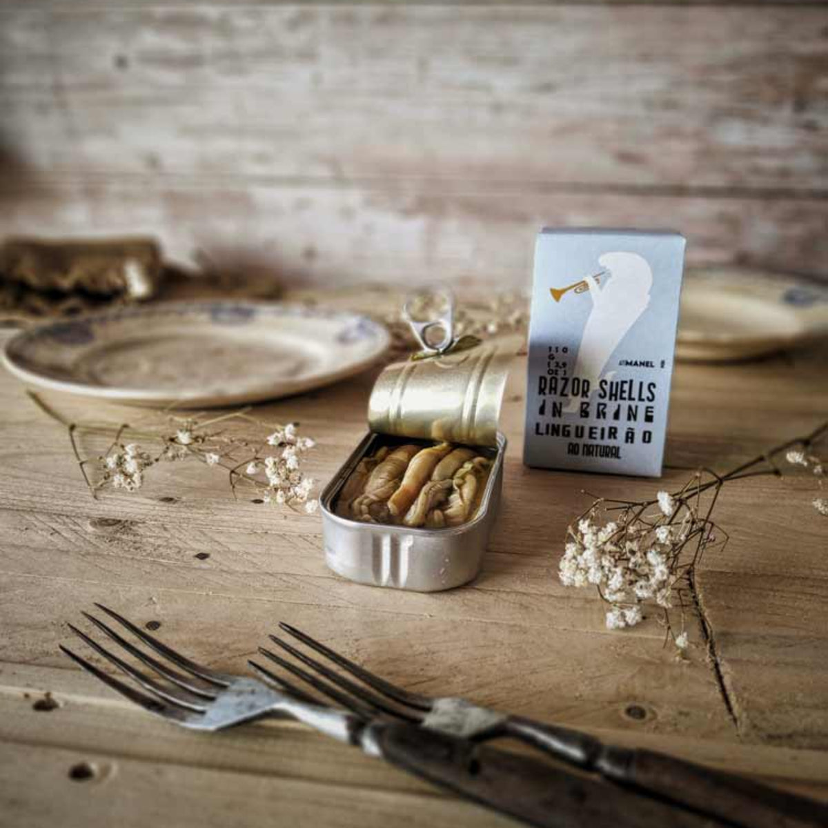 A styled tabletop scene with a small open metal tin of food, a printed card or box standing beside it, plates, dried flowers, and cutlery on a rustic wooden surface.