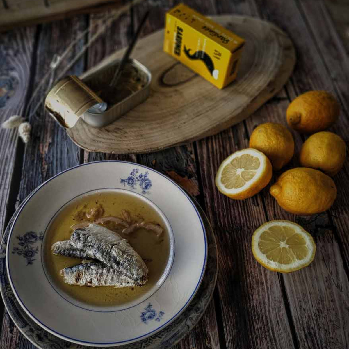Dish of fish with a bowl of soup on a wooden table with lemons and a box of matches.
