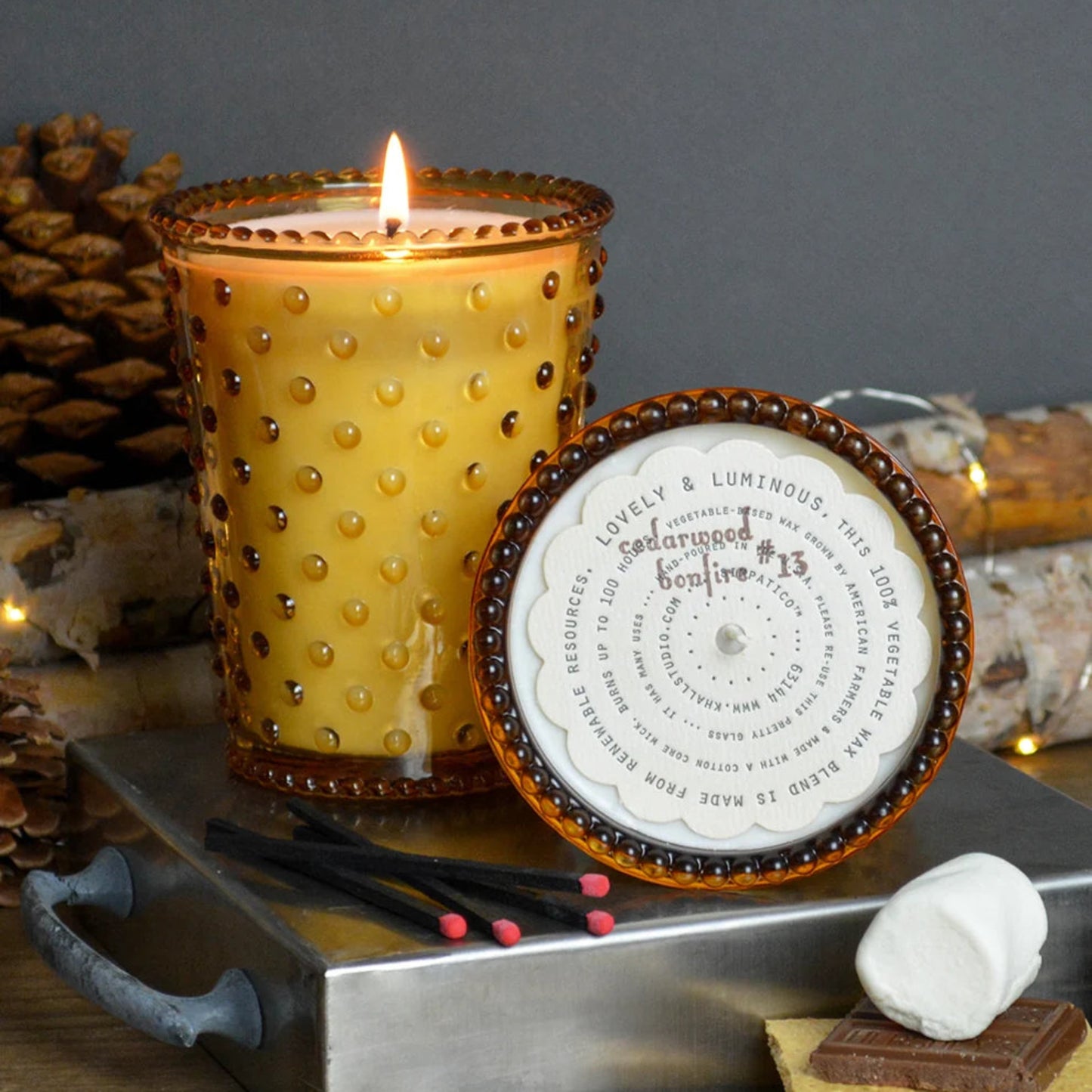 A lit amber glass candle with a textured bubble pattern sits on a metal tray beside its printed lid. The scene includes s'mores props, pinecones, wood logs, and a dark gray background.