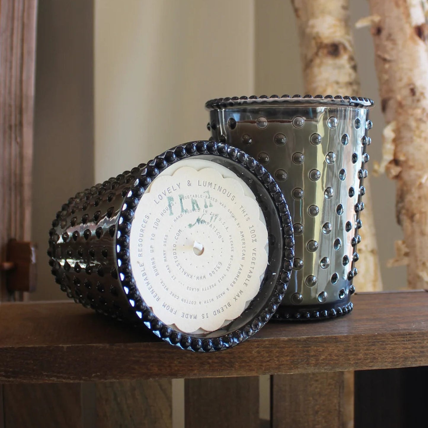 Two decorative candle holders with a dark metallic, beaded, and perforated design, one standing upright and the other lying on its side to show the wax label, displayed on a wooden shelf.
