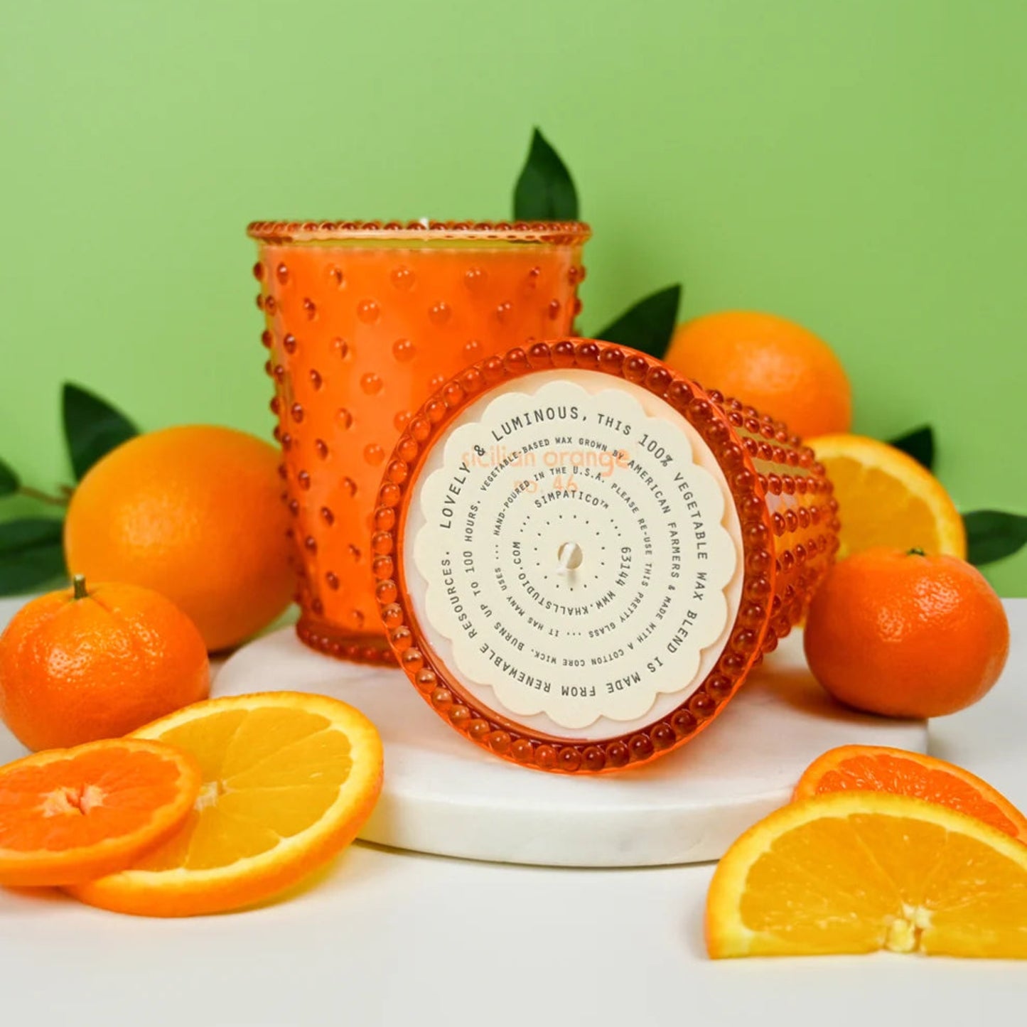 Orange textured candle with a label surrounded by oranges on a green background