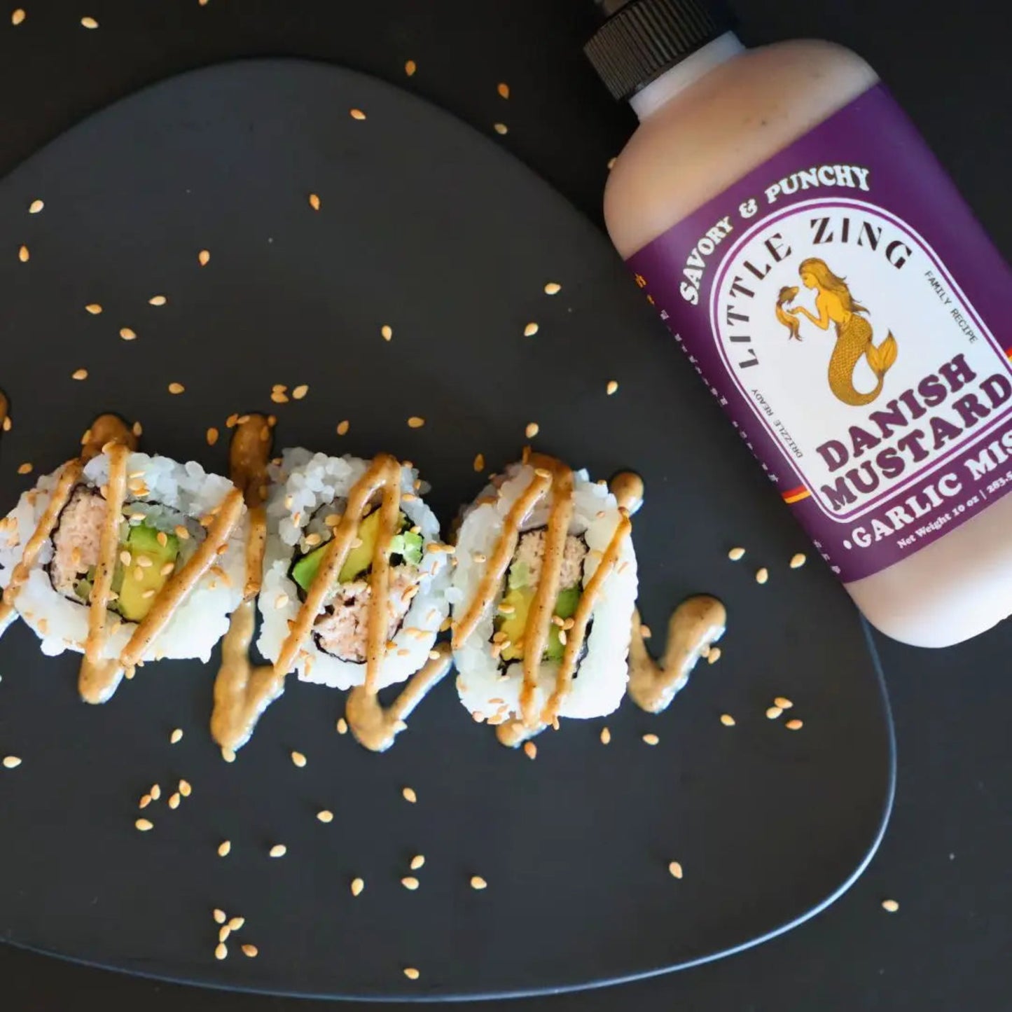 Sushi rolls with a drizzle of mustard on a black plate, accompanied by a bottle of Little Zing Danish Mustard.