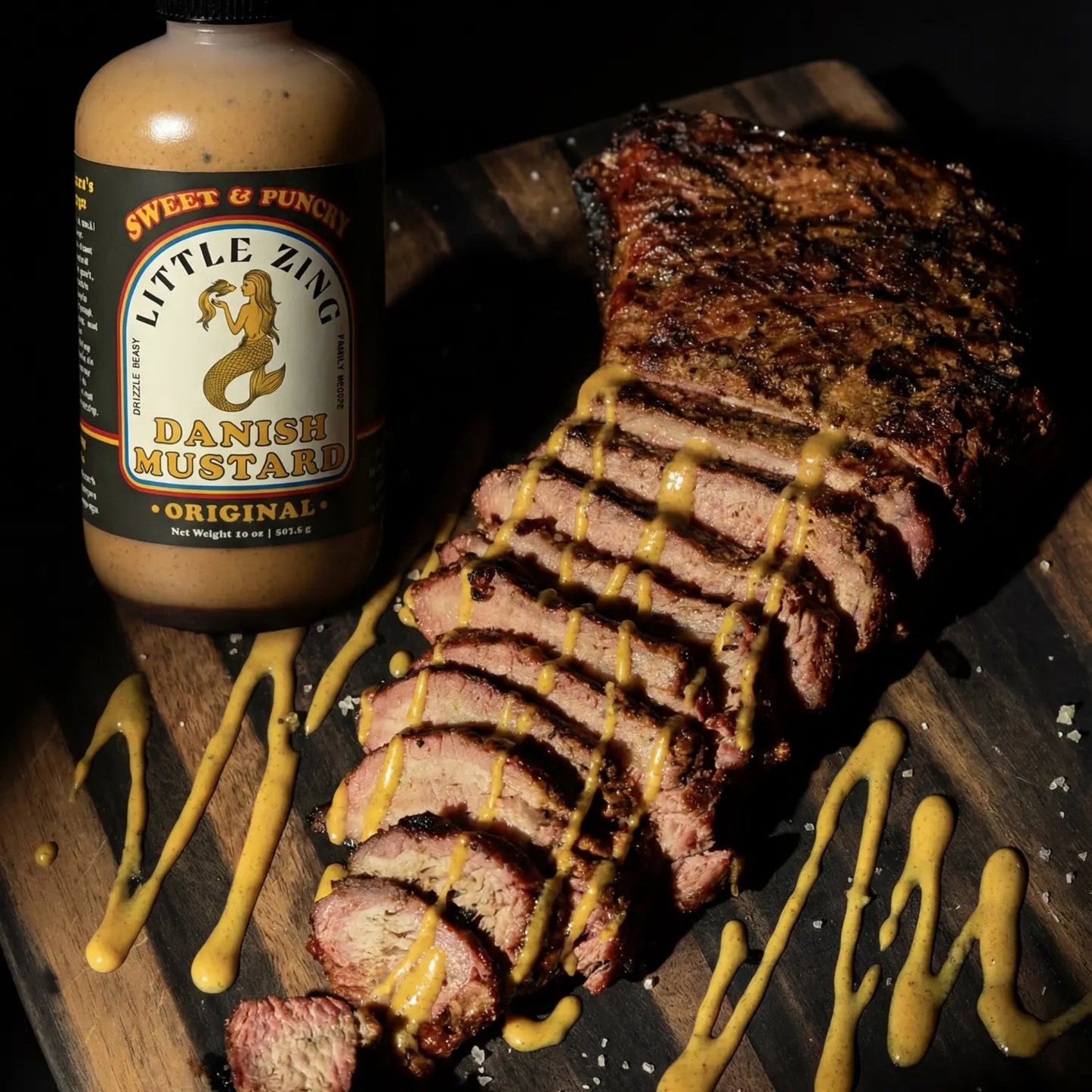 Sliced meat with mustard drizzle next to a bottle of Little Zing Danish Mustard on a wooden surface.