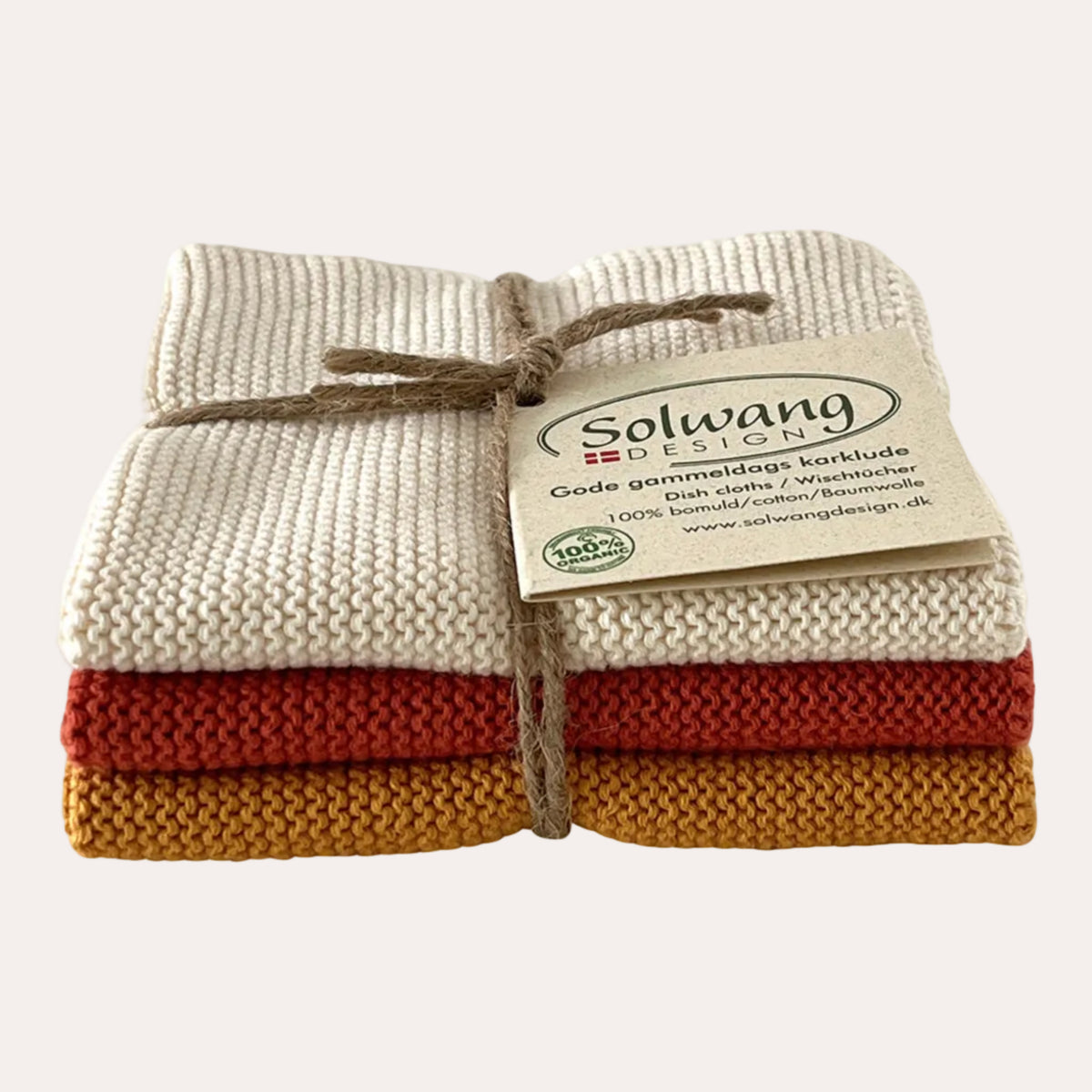 A stack of three knitted wool washcloths in cream, rust red, and mustard yellow, tied together with a twine bow and a paper brand tag reading Solwang Design.