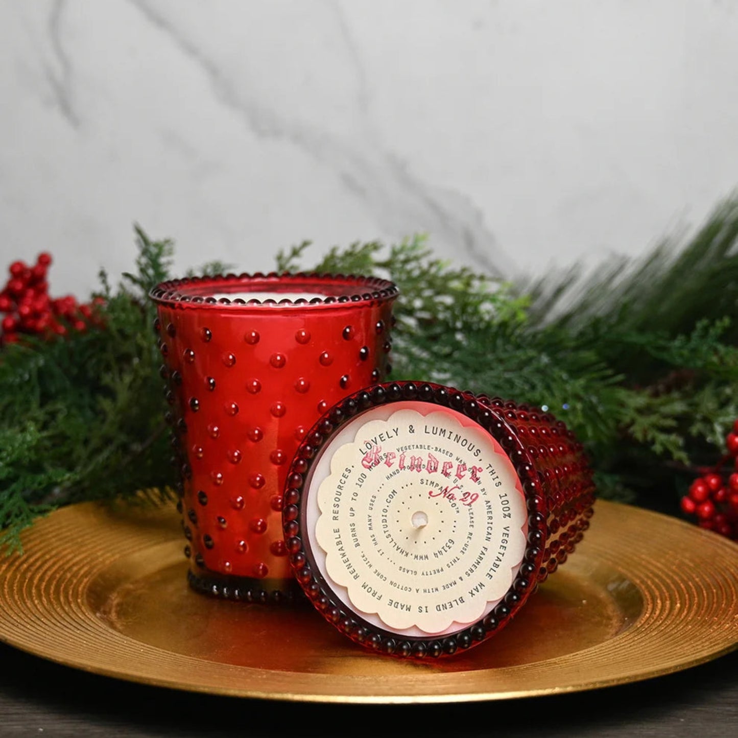 A red textured glass candle holder with a matching lid, displayed on a gold plate with green pine branches and red berries in the background.