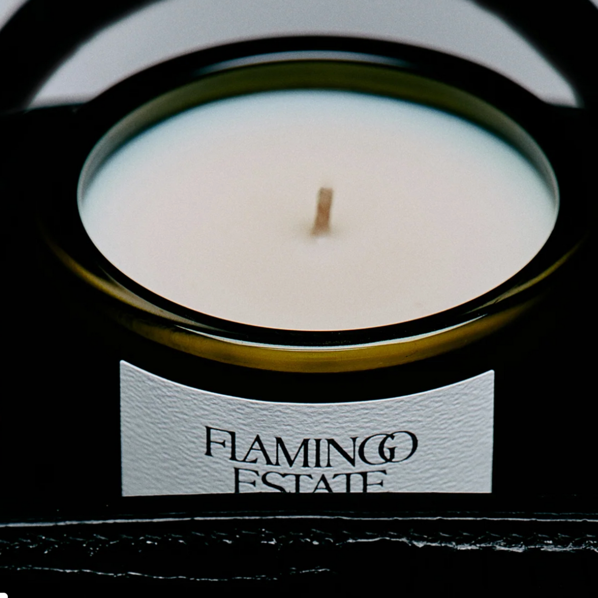 A close-up of a black glass jar candle with a white wax surface and a single wick, featuring a white label that reads 'FLAMINGO ESTATE' on a dark background.