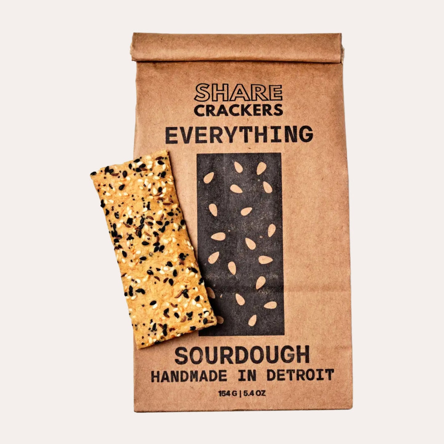 A single slice of Share Crackers Everything Sourdough Handmade in Detroit cracker, topped with seeds, shown next to its brown kraft paper packaging bag on a light beige background.