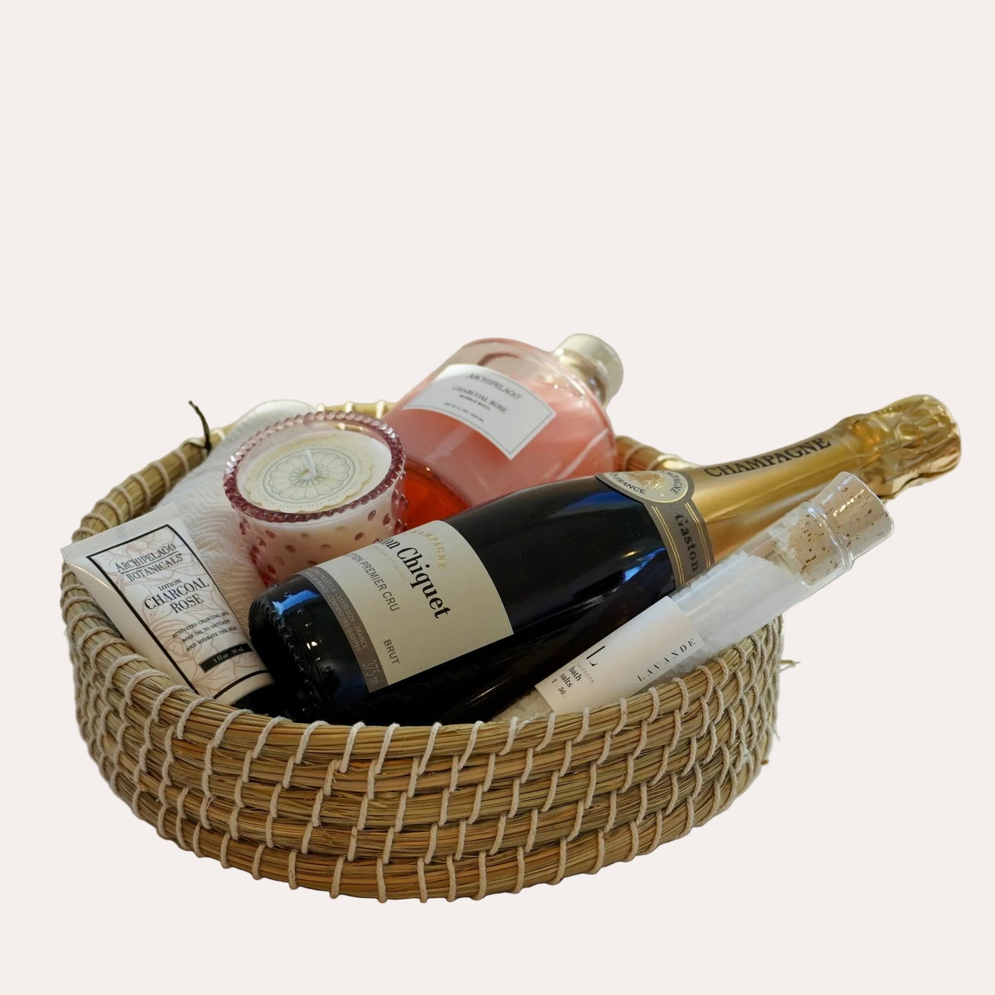 Bottle of champagne and bath products in a woven basket on a white surface