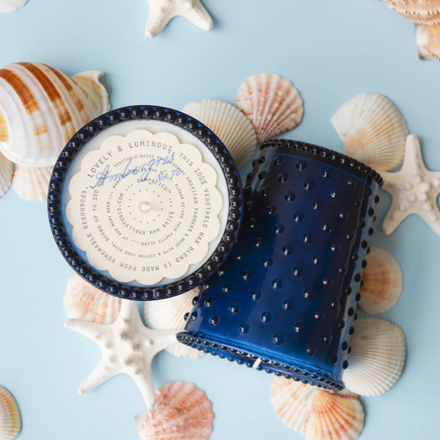 A round blue candle with its lid off, showing the printed label, styled on a light blue background with seashells and starfish.